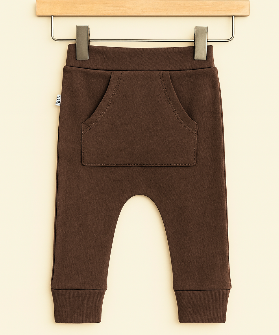 Calça de bebê marrom com bolso canguru em algodão premium, usada por bebê de 9 meses em ambiente boho chic com luz natural, madeira e plantas