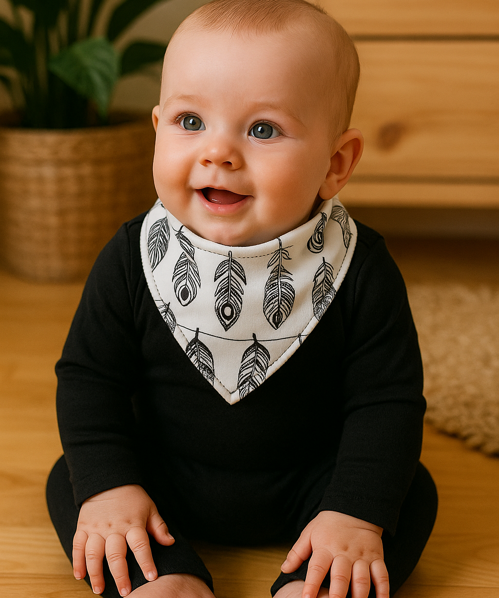 Bebê de 6 meses usando bandana babador com estampa de penas pretas em fundo branco, body preto de manga longa, sentado em ambiente acolhedor com madeira pinus e plantas naturais.

