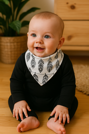 Bebê de 6 meses usando bandana babador com estampa de penas pretas em fundo branco, body preto de manga longa, sentado em ambiente acolhedor com madeira pinus e plantas naturais.

