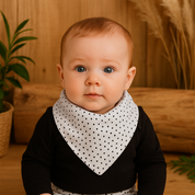 Bebê de 4 meses com olhos claros e cabelo castanho claro, usando bandana babador estampa poá e conjunto coordenado com body preto e calça branca com bolinhas pretas, sentado em ambiente com madeira pinus, folhas verdes e luz natural suave.

