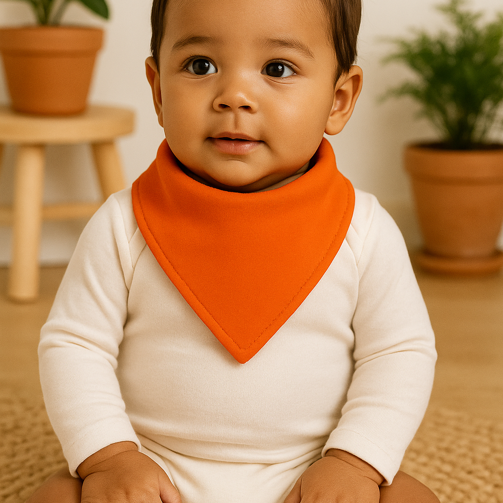 Bebê de pele morena usando bandana babador laranja e body neutro de manga longa, sentado em ambiente com elementos naturais, madeira clara e plantas ao fundo.

