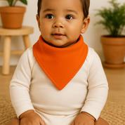 Bebê de pele morena usando bandana babador laranja e body neutro de manga longa, sentado em ambiente com elementos naturais, madeira clara e plantas ao fundo.

