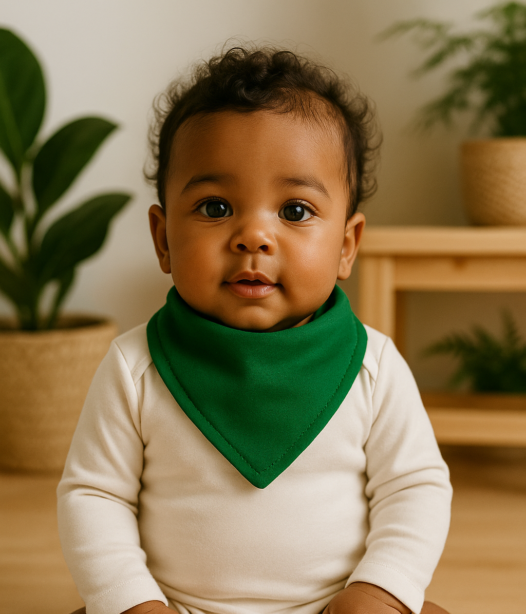 Bebê de pele morena e cabelo crespo usando bandana babador verde bandeira e body neutro manga longa, sentado em ambiente iluminado com piso de madeira clara, plantas naturais e móveis em pinus.

