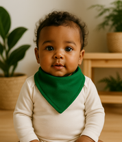 Bebê de pele morena e cabelo crespo usando bandana babador verde bandeira e body neutro manga longa, sentado em ambiente iluminado com piso de madeira clara, plantas naturais e móveis em pinus.

