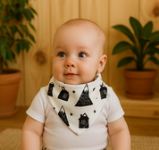 Bebê de cerca de 6 meses usando bandana babador com estampa de casinhas nórdicas, body branco e calça combinando, sentado em ambiente acolhedor com madeira pinus e plantas naturais ao fundo.

