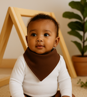 Bebê de 6 meses usando bandana babador lisa marrom café e body branco de manga longa, sentado em ambiente com tapete claro, móveis em madeira pinus e plantas naturais.


