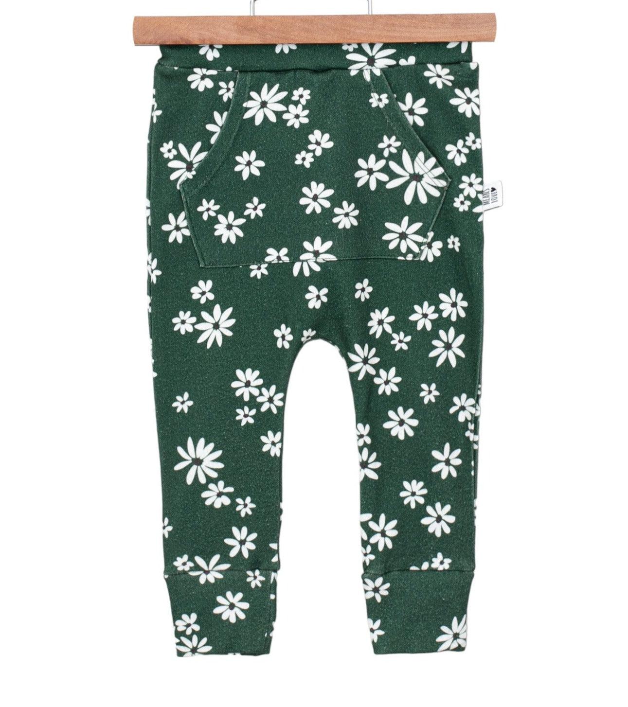 Bebê pequeno de pé sobre tapete de linho bege, usando calça verde com estampa floral branca da coleção Daisy da Means Love Baby.

