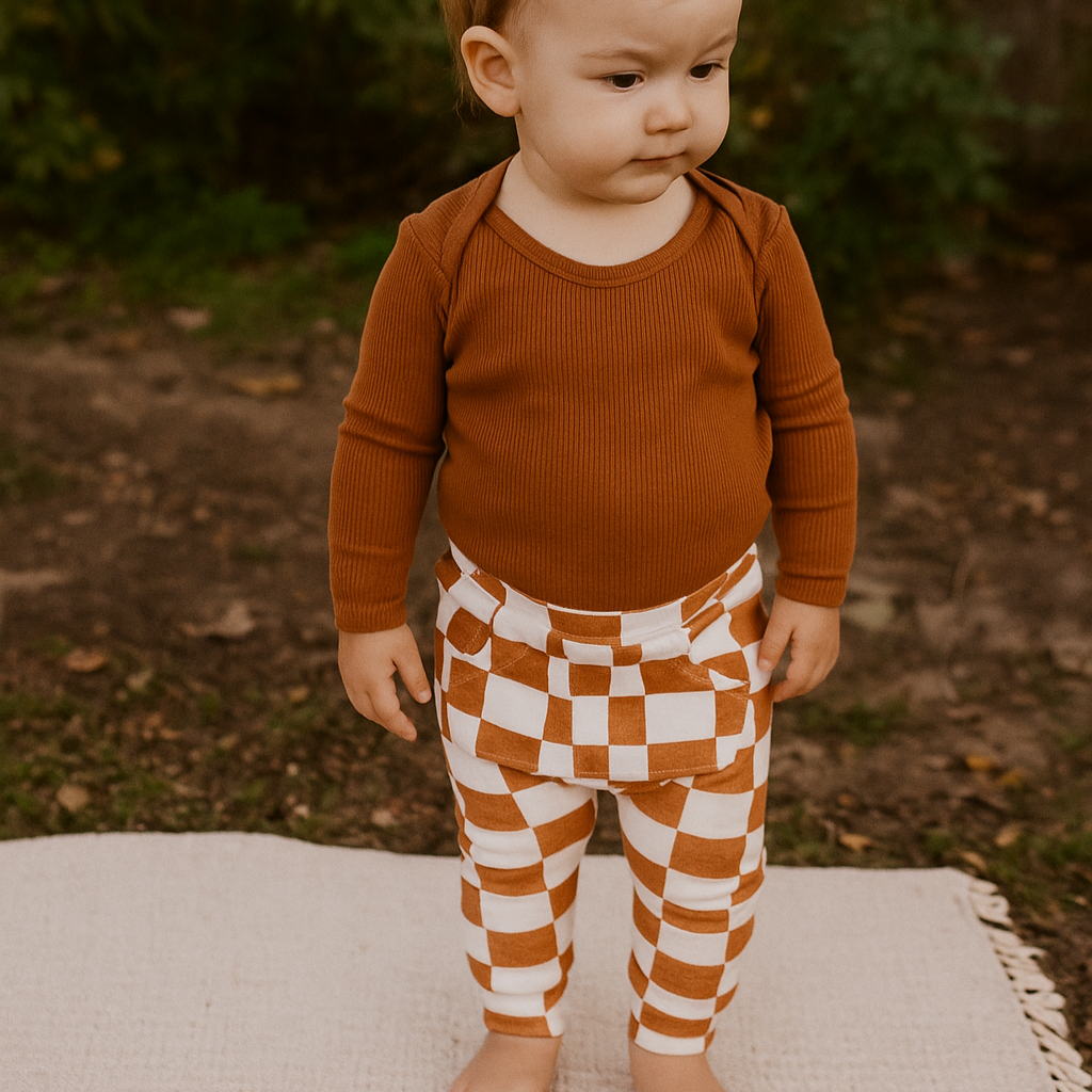 Bebê pequeno de pé sobre fundo de linho bege, usando body canelado mostarda e calça infantil com estampa quadriculada terracota da coleção Grid da Means Love Baby.