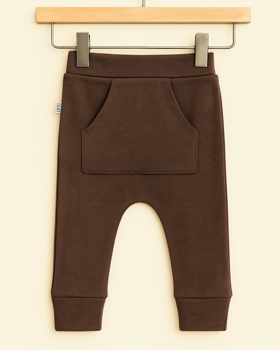 Calça de bebê marrom com bolso canguru em algodão premium, usada por bebê de 9 meses em ambiente boho chic com luz natural, madeira e plantas


