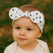 Bebê de 1 ano ao ar livre usando laço de cabelo infantil branco com estampa de gotas pretas e body bege.