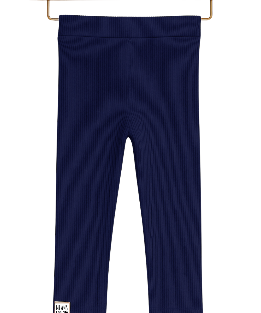 Legging infantil canelada azul marinho em algodão premium, com cós anatômico e modelagem confortável, pendurada em cabide de madeira sobre fundo neutro boho.

