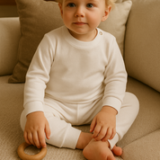 Bebê de 4 meses usando pijama branco canelado de duas peças em ambiente boho chic com luz natural e elementos como madeira e plantas.

